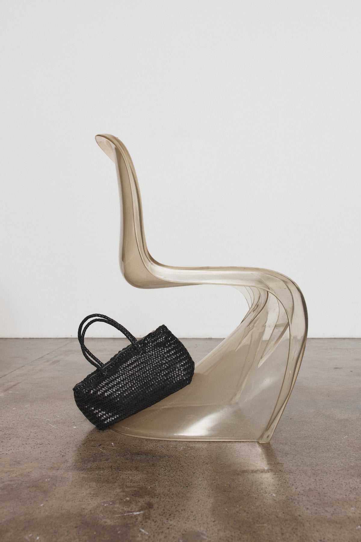 A black woven leather handbag with double top handles rests inside a sculptural, transparent beige plastic chair on a concrete floor against a white wall.