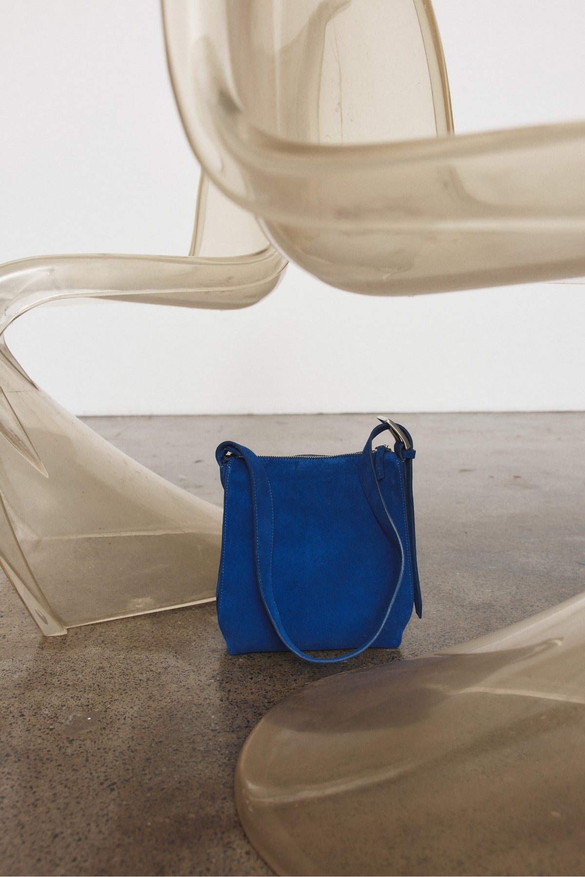 A bright blue suede bucket bag with a matching shoulder strap, placed on a concrete floor between translucent modern chairs.